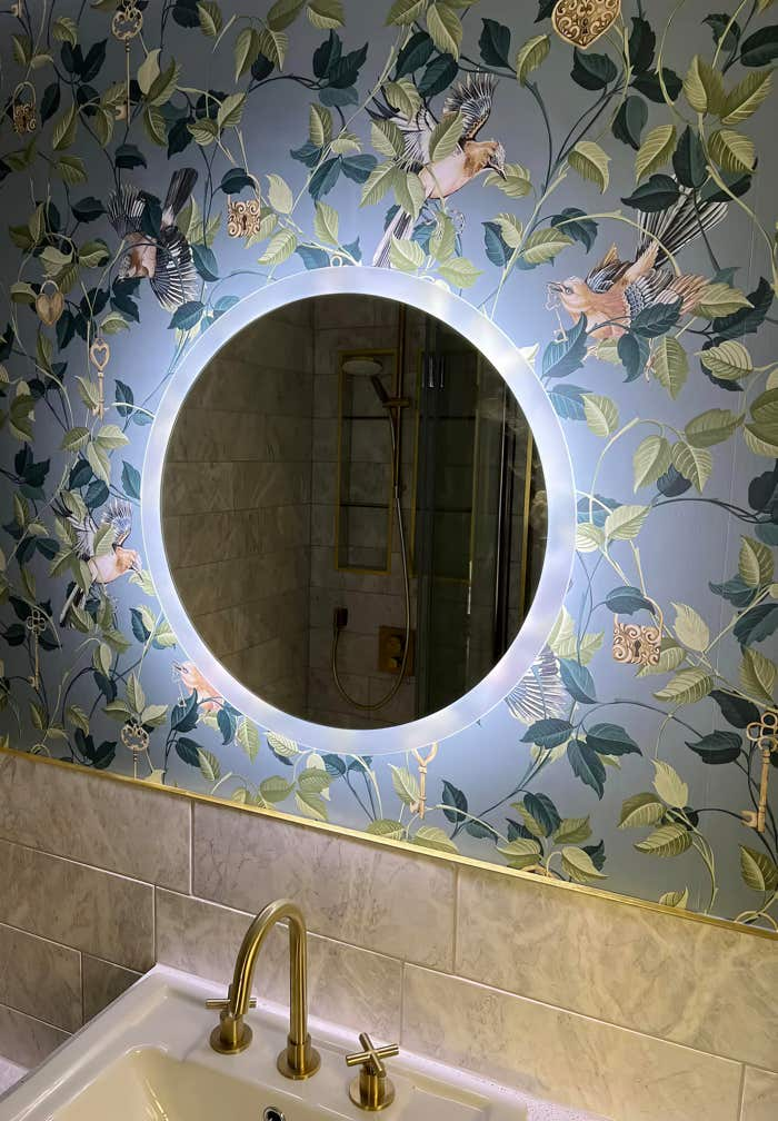 Image of LED illuminated circular mirror above a sink with floral wallpaper and polished gold accessories.